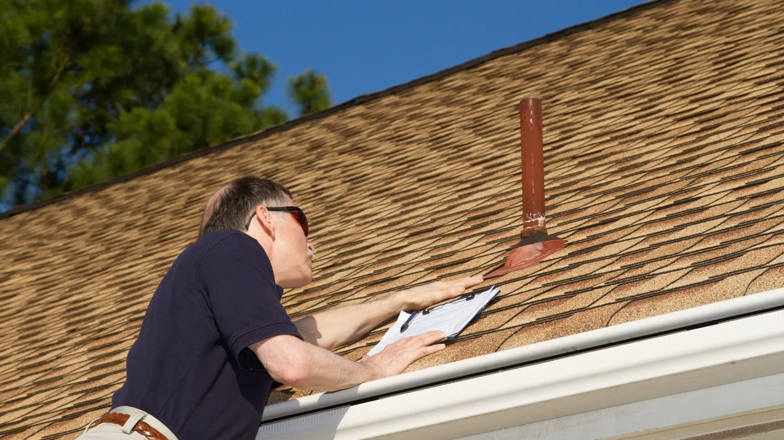 An inspector checking shingles around a vent pipe with a clipboard